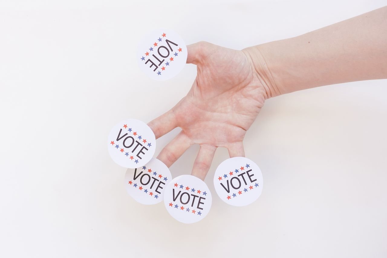 A hand displays several 'Vote' stickers on a white background, symbolizing political participation.